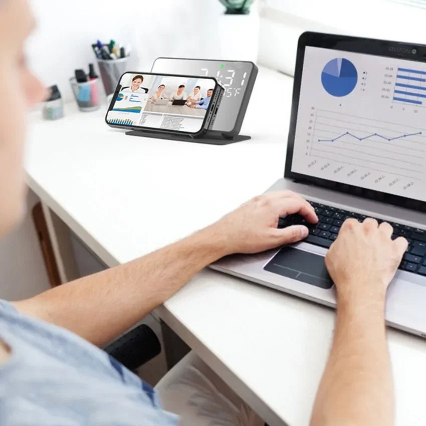 Person using a laptop with a smartphone on a stand displaying a video call, in a home office setting.