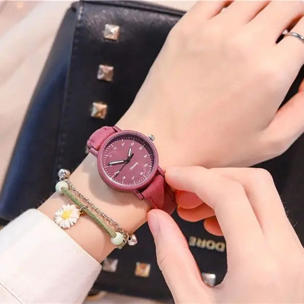 Person wearing a maroon wristwatch with a matching strap and daisy charm bracelet, adjusting the watch, with a black studded handbag in the background.