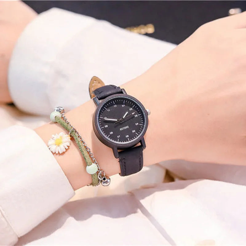 Black wristwatch with a black leather strap on a wrist, paired with a daisy charm bracelet.
