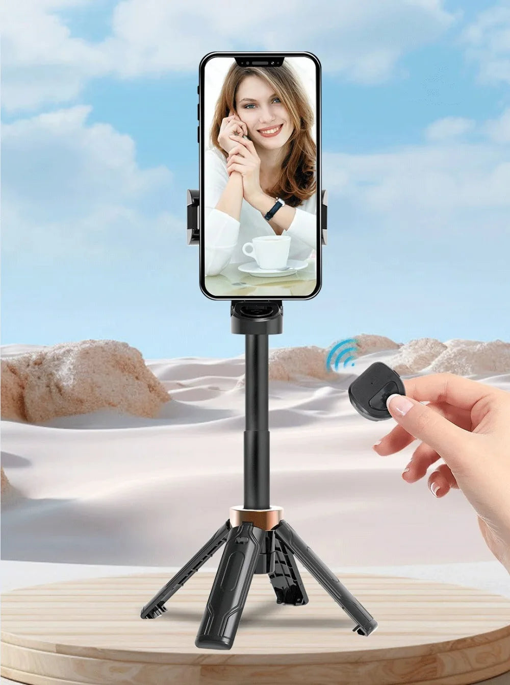 Smartphone with a screen displaying a woman's face, placed on a tripod with a hand holding a remote control against a desert landscape.