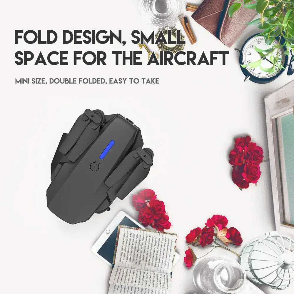 E88 Pro foldable drone with blue indicator light on a white surface alongside flowers and stationery items