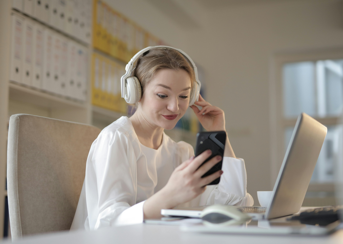 Smiling woman wearing white wireless headphones using a smartphone while sitting at a desk with a laptop