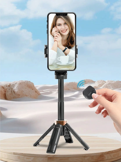 Smartphone with a screen displaying a woman's face, placed on a tripod with a hand holding a remote control against a desert landscape.
