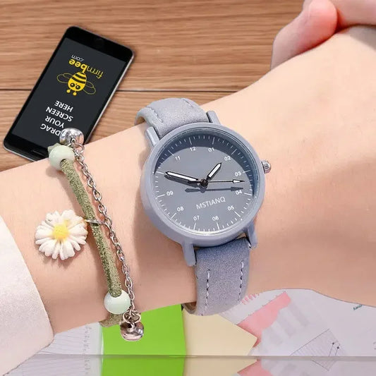 Light blue wristwatch with a minimalist dial, worn on a wrist alongside a daisy charm bracelet, with a smartphone in the background.